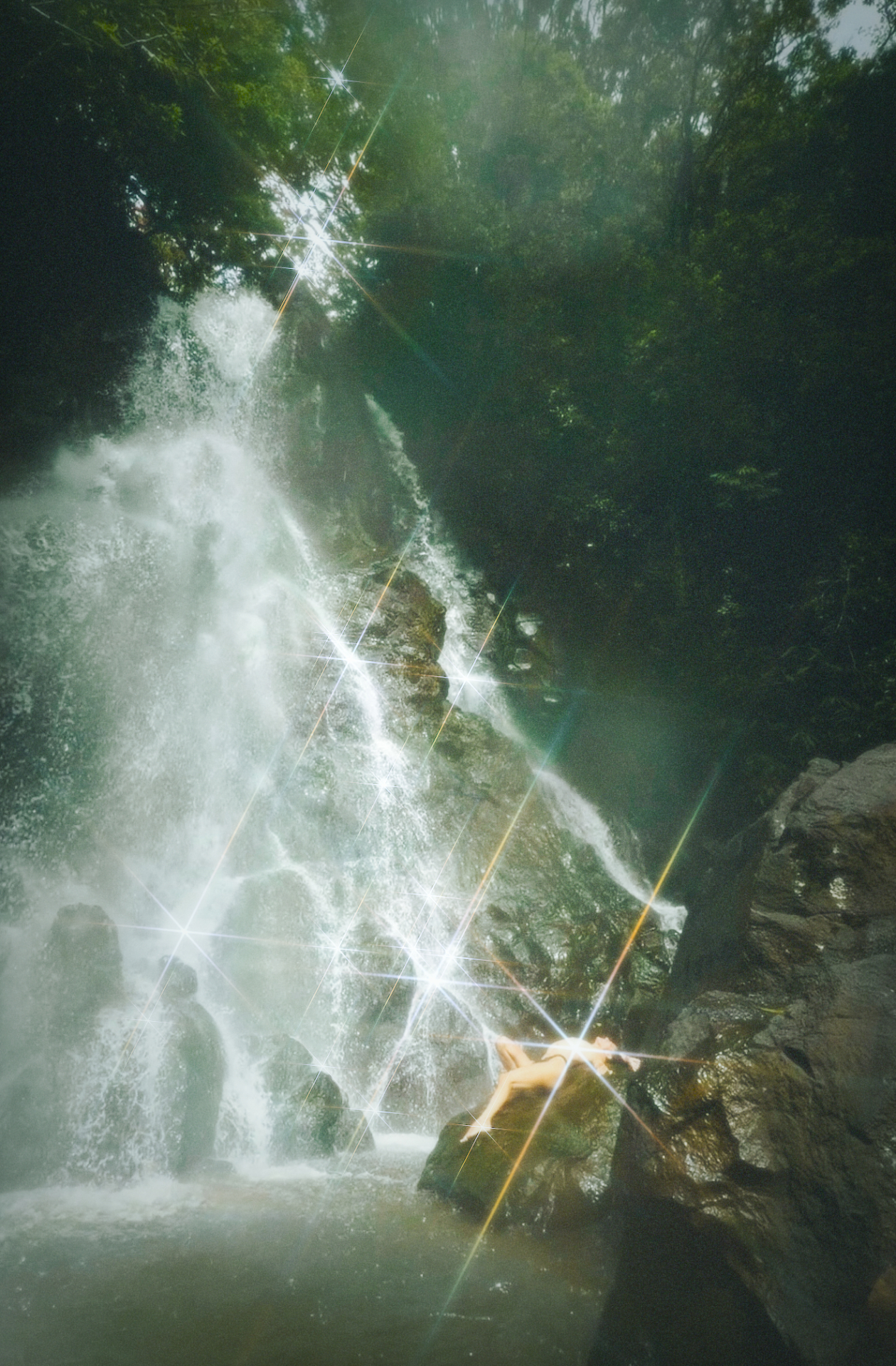 A figure reclining on sunlit rocks beside a waterfall — lens flares cutting through the mist.